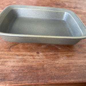Puede incluir: Una fuente de horno rectangular de cerámica verde oliva con esquinas redondeadas. La fuente está colocada sobre una superficie de madera. La fuente para hornear está vacía. La fuente mide unos 25 cm de largo y 20 cm de ancho.