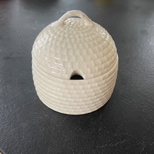 Könnte beinhalten: Weiße Keramik-Honigtopf mit einem Deckel in Form eines Bienenstocks. Der Topf hat eine kleine Öffnung an der Seite zum Ausgießen von Honig.