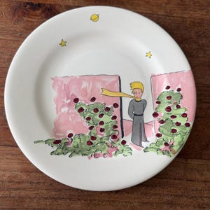 Op de afbeelding: Een wit keramieken bord met een kleurrijke illustratie van een jong persoon met blond haar en een gele sjaal, staand tussen twee roze muren. Er zijn groene rozenstruiken met rode bloemen afgebeeld, samen met gele sterren en een planeet.