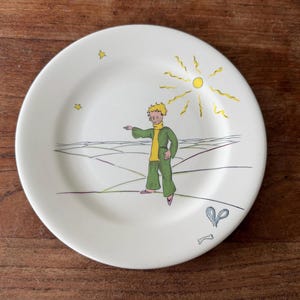 May include: A white ceramic plate featuring a colourful illustration of a person with blonde hair wearing a green jacket and yellow shirt. The scene includes a yellow sun, two small stars, a landscape with lines, a small grey heart, and a bone.