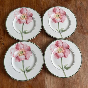 Może przedstawiać: Cztery białe ceramiczne talerze z różowym wzorem kwiatowym. Każdy talerz ma duży różowy kwiat z zieloną łodygą i liśćmi. Talerze mają zieloną obwódkę i są ułożone na drewnianej powierzchni.