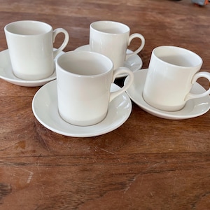 May include: Four white ceramic coffee cups with matching saucers. The cups and saucers are arranged in a row on a wooden table.