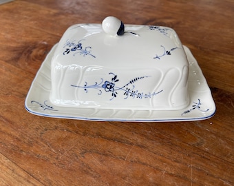 May include: White ceramic butter dish with a lid, decorated with blue floral patterns. The dish is rectangular with a scalloped edge and a small, round knob on top. The base is also rectangular with a blue trim.