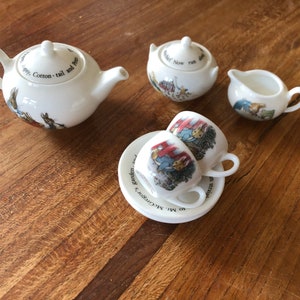May include: A miniature white porcelain tea set featuring Peter Rabbit illustrations and text. The set includes a teapot with a lid, a lidded sugar bowl, a creamer, and two teacups resting on a saucer. Each piece displays colourful character scenes.