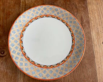 Op de afbeelding: Een ronde keramische plaat met een wit midden en een patroonrand. De rand heeft een lichtbeige achtergrond met blauwe ruitvormen en een oranje en groen fruitontwerp. De plaat staat op een houten oppervlak.