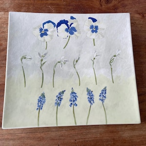 Puede incluir: Plato cuadrado de cerámica con un diseño floral pintado a mano. El plato presenta pensamientos blancos y azules, campanillas de invierno y jacintos de uva azules. El fondo es de un suave verde amarillento.