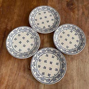 Op de afbeelding: Vier crèmekleurige keramische borden met een blauw bloemmotief en een decoratieve blauwe rand. De borden zijn gerangschikt op een houten oppervlak. De borden zijn een set servies.
