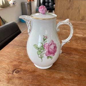 Puede incluir: Crema de porcelana blanca con ribete dorado y diseño de rosa rosa. La crema tiene una tapa con una rosa rosa en la parte superior.