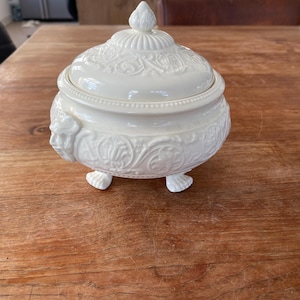 Puede incluir: Azucarero de cerámica blanca con tapa y diseño floral ornamentado. El tazón tiene tres patas y una cabeza de león a cada lado.