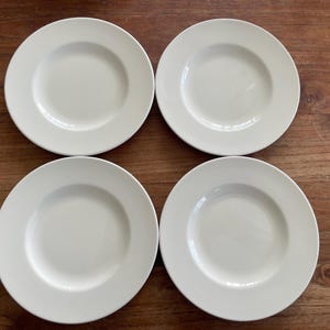 Puede incluir: Cuatro platos de cena de cerámica blanca con un borde ligeramente elevado. Los platos están dispuestos en forma de cuadrado sobre una superficie de madera.