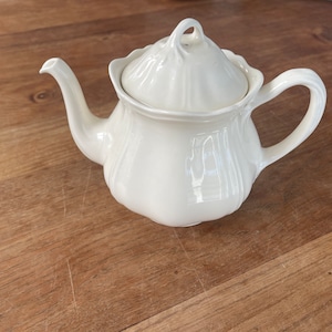 Puede incluir: Tetera de cerámica blanca con tapa y asa. La tetera tiene un diseño clásico con un cuerpo redondeado y un pico.
