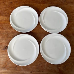 Op de afbeelding: Vier witte keramische borden met een spiraalvormig ontwerp. De borden zijn in een vierkant op een houten oppervlak gerangschikt. De borden hebben een gladde, glanzende afwerking en een licht verhoogde rand.