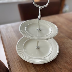 May include: A two-tiered serving stand with a silver handle and two off-white plates. The plates have a ridged border design. The stand is on a wooden table. The handle is a loop shape.