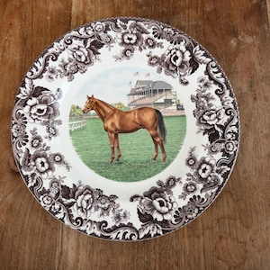 Puede incluir: Un plato redondo de cerámica con una ilustración central de un caballo marrón de pie en un campo verde frente a una tribuna. El borde está decorado con un patrón floral en marrón oscuro y blanco con hojas y flores entrelazadas.