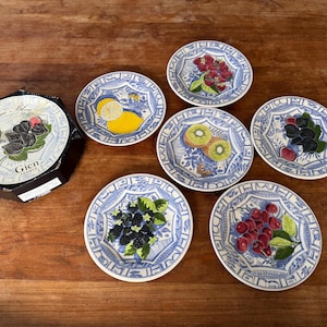 May include: A set of six decorative ceramic plates with blue and white patterns, each featuring a different fruit design: lemon, kiwi, raspberries, blackberries, cherries, and mulberries. A box of fig-flavored treats is also present.