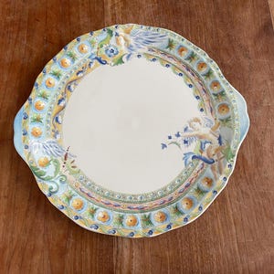 Peut inclure: Un plat de service en céramique blanche avec un bord festonné. Le plat est décoré d'un motif floral bleu, jaune et vert et d'une figure de chérubin.