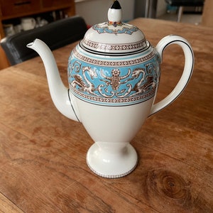 Könnte beinhalten: Eine weiße Teekanne aus Keramik mit einem blauen und braunen Blumendesign. Die Teekanne hat einen Deckel mit einem goldenen Knopf und einen Ausguss an der Seite. Die Teekanne steht auf einem Holztisch.