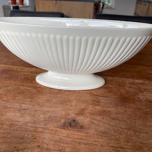 Puede incluir: Un cuenco de cerámica blanca con un diseño acanalado y una base de pedestal. El cuenco está sobre una superficie de madera.