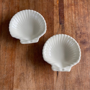 Op de afbeelding: Twee witte, schelpvormige schalen van keramiek. De schalen zijn glad en hebben een glanzende afwerking.