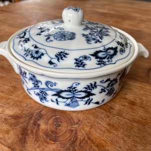 Puede incluir: Una cacerola de porcelana blanca con un patrón floral azul y blanco. La cacerola tiene una tapa con un pomo en la parte superior.