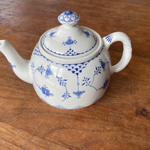 Puede incluir: Tetera de porcelana blanca con un estampado floral azul. La tetera tiene un pico curvo, un cuerpo redondeado y una tapa con un pomo decorativo. El asa también es curva. El diseño floral azul se repite en la tapa y el cuerpo.