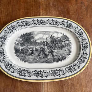 Puede incluir: Plato ovalado para servir con una escena pastoral en blanco y negro en el centro. El borde presenta un diseño floral en blanco y negro, con un ribete amarillo. El plato está sobre una superficie de madera.