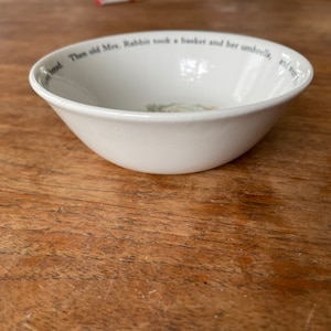 Może przedstawiać: Biała ceramiczna miska z czarnym rantem i tekstem "Then old Mrs. Rabbit took a basket and her umbrella, and went to", klasyczna ilustracja z książki dla dzieci.
