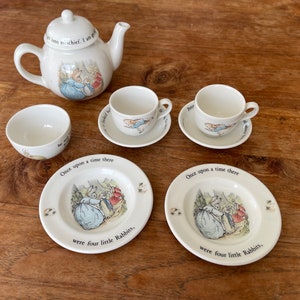 Può includere: Un set da tè per bambini bianco con illustrazioni di Peter Rabbit. Il set include una teiera, due tazze da tè, due piattini, una ciotola e due piatti. I piatti hanno il testo "C'era una volta, c'erano quattro coniglietti." stampato su di essi.