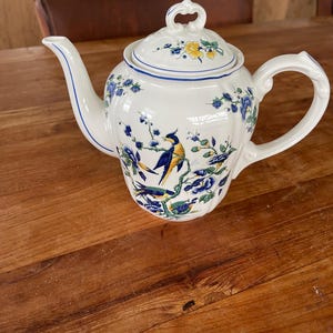 Può includere: Una teiera in ceramica bianca con un motivo floreale blu e giallo e un motivo di uccello. La teiera ha un coperchio con un pomo in cima.