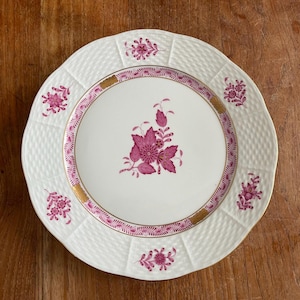 May include: White ceramic plate with a scalloped edge and a woven texture. The plate features a central floral design and a pink and gold border. Additional pink floral accents are placed around the rim.