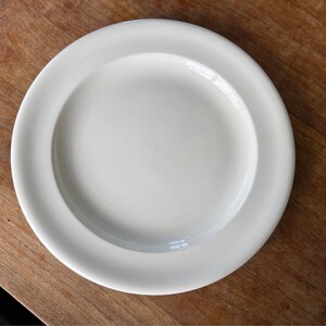 Puede incluir: Un plato de cerámica blanco con un borde ligeramente elevado. El plato está vacío y tiene un acabado liso y brillante.