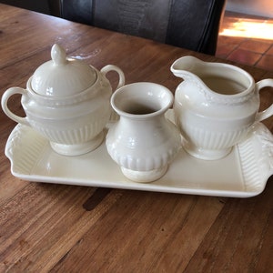 Può includere: Un servizio da tè color crema su un vassoio rettangolare. Il set include una zuccheriera con coperchio, una lattiera e un piccolo vaso. I pezzi hanno un design a coste e sono posizionati su un vassoio con un bordo a forma di foglia. Il set è su un tavolo di legno.