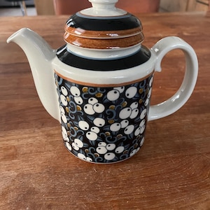 Puede incluir: Una tetera de cerámica blanca con una tapa negra y marrón. La tetera tiene un patrón floral blanco y negro con puntos blancos.