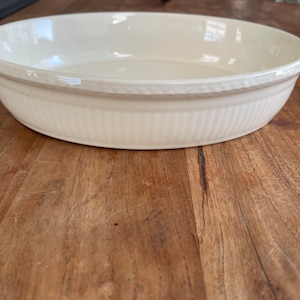 Può includere: Pirofila rotonda in ceramica color crema, con bordo strutturato e lati scanalati verticali. Il piatto è su una superficie di legno. Adatta per cuocere al forno e servire.