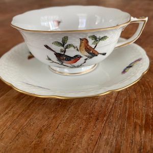 Op de afbeelding: Een wit porseleinen theekopje en schotelset met een gouden rand. Het kopje en de schotel hebben een delicaat bloemenmotief met twee vogels die op een tak zitten.