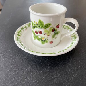 Pode incluir: Uma xícara de café e um pires de cerâmica branca com um padrão de morangos verdes e vermelhos.