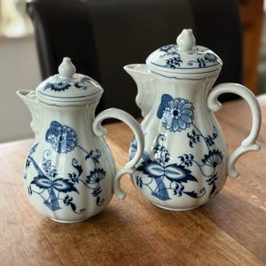 May include: Two white ceramic jugs with blue floral patterns and matching lids. The jugs have curved handles and spouts. The design features stylized flowers and leaves. They are placed on a wooden surface.