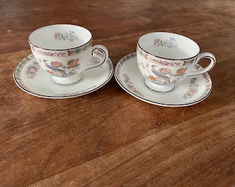 May include: Two white teacups and saucers with a floral and bird design. The cups and saucers have a black trim. The teacups have a handle and sit on a wooden surface.