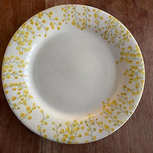 May include: A white ceramic plate with a yellow floral pattern around the rim. The design features small yellow flower clusters on thin green stems. The plate has a simple, elegant design, suitable for serving food.