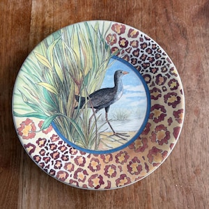 May include: A decorative plate featuring a bird in a marsh scene, surrounded by tall green and yellow reeds. The plate's border has a leopard print design in shades of brown and tan. The bird is depicted with gray and black feathers.