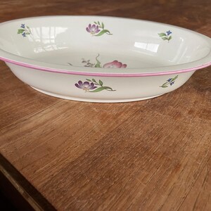 May include: Oval, white ceramic serving dish with a pink rim. The dish is decorated with floral designs in purple, pink, and blue. The dish is sitting on a wooden surface.