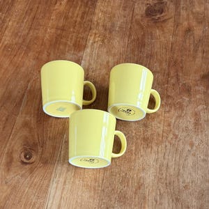 Op de afbeelding: Drie gele keramische mokken met gebogen handvatten op een houten oppervlak. De mokken hebben een eenvoudige, cilindrische vorm en een heldere, effen gele kleur. De bodems van de mokken hebben een witte rand en een gedrukt logo.