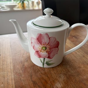 Op de afbeelding: Witte keramische theepot met een groot roze bloemdesign. De theepot heeft een gebogen tuit, een afgeronde handgreep en een deksel met een kleine knop. Een groene band accentueert het deksel. De theepot staat op een houten oppervlak.