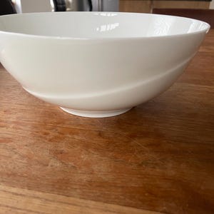 以下が含まれることがあります： 波打つデザインの白いセラミックボウル。ボウルは木製のテーブルの上に置かれています。