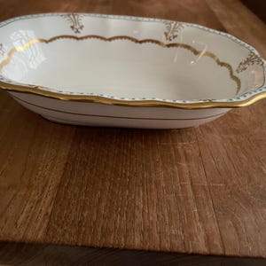 May include: White oval serving bowl with a gold and blue trim. The bowl has a scalloped edge and a decorative design.