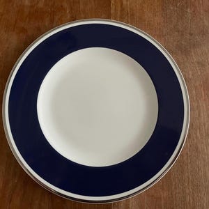 Puede incluir: Un plato de cerámica blanco con un borde azul y un ribete plateado. El plato tiene un diseño sencillo y es perfecto para servir una variedad de platos.