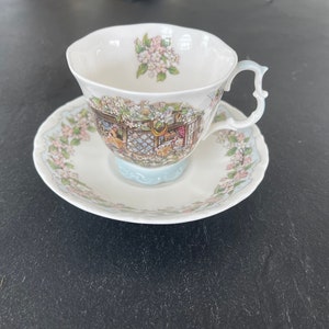 May include: A white teacup and saucer with a floral pattern and a scene of a cottage with animals. The cup has a delicate handle and the saucer has a scalloped edge.