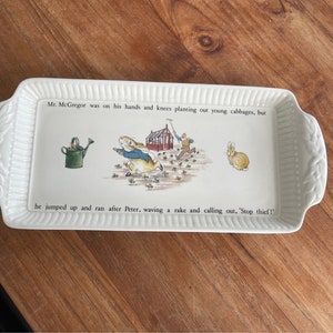 Puede incluir: Bandeja de servir de cerámica blanca con una ilustración de Peter Rabbit. La bandeja presenta una escena de Mr. McGregor haciendo jardinería y Peter Rabbit huyendo. El texto en la bandeja dice "Mr. McGregor estaba de rodillas plantando coles jóvenes, pero saltó y corrió tras Peter, agitando un rastrillo y gritando: '¡Alto, ladrón!'".