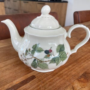 Può includere: Una teiera in porcellana bianca con un design floreale con foglie verdi e more. La teiera ha un coperchio e un manico.