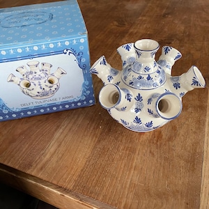 Könnte beinhalten: Eine weiß-blaue Delft-Tulpenvase mit sieben Armen, neben der passenden blauen Box. Die Vase hat ein traditionelles niederländisches Design mit floralen Mustern. Auf der Box steht "DELFT TULIPVASE 7 ARMS".